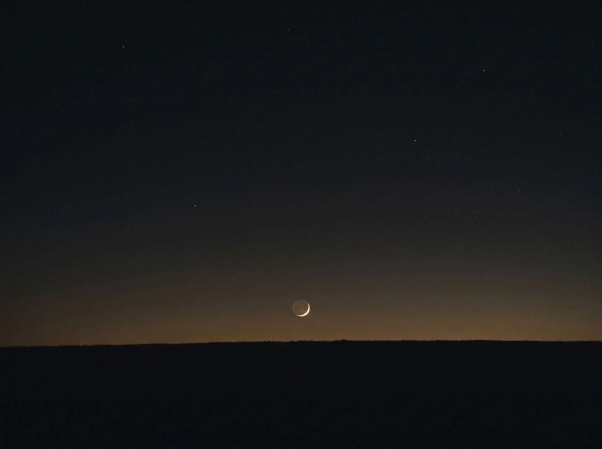 Una luna creciente muy fina sobre un horizonte oscuro, en el último minuto de luz del día.